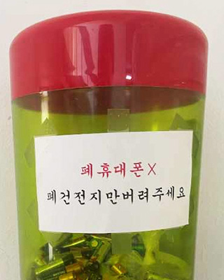 노란색 불투명한 몸통에 윗부분엔 빨간색의 플라스틱 길죽한 통의 수거함 모습, 폐휴대폰x 폐건전지만 버려주세요 라는 문구가 적혀있다