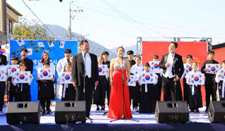 무대위 빨간드레스 입은 여자가 서있고 뒷편에는 청년과 어린이들이 태극기를 를고 있는 모습 