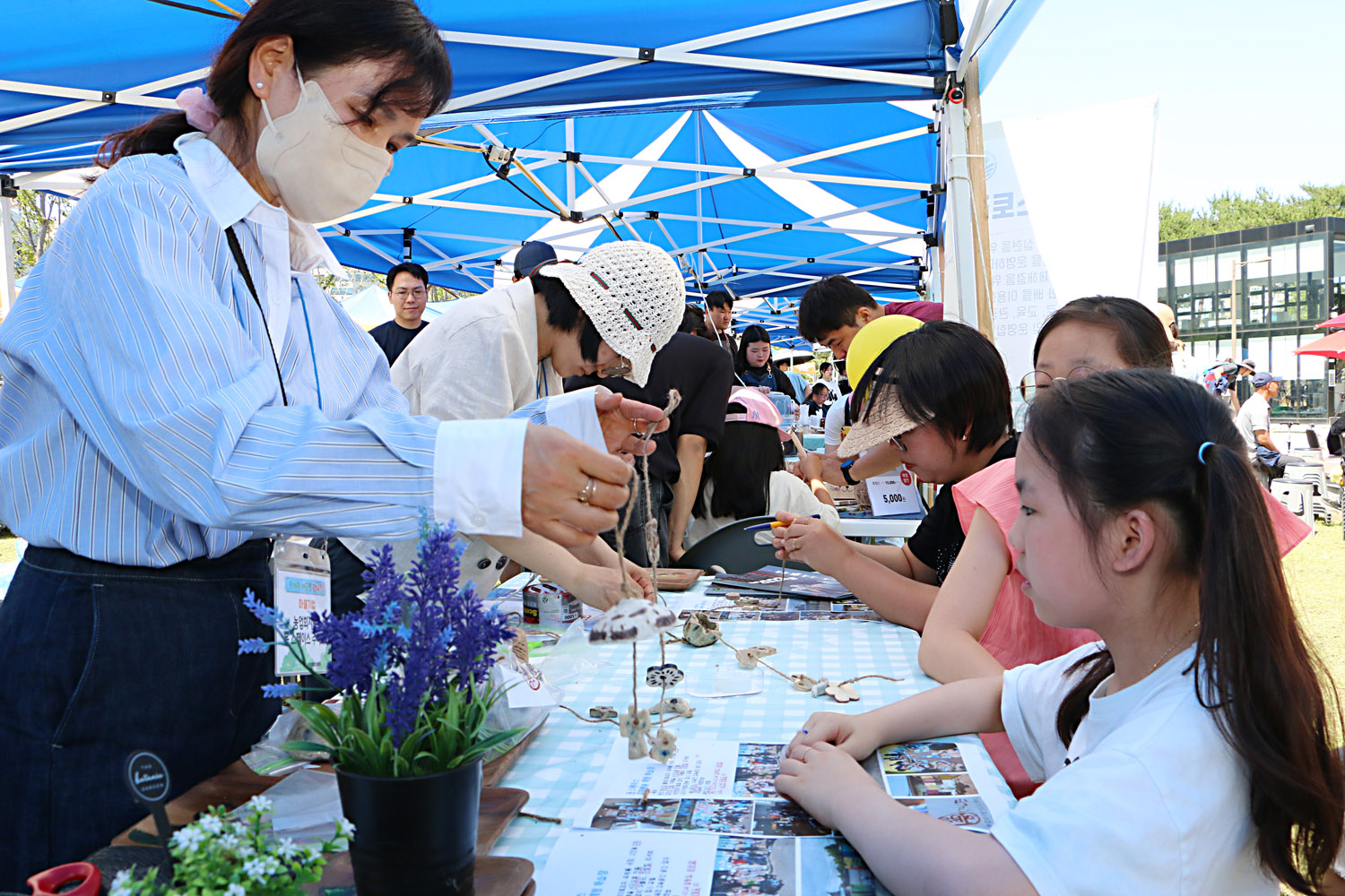 多가치 어울림 한마당 기념식 행사장 부스에서 목걸이 공예품을 만들어 건네는 관계자와 그것을 바라보고 있는 여자 아이의 모습