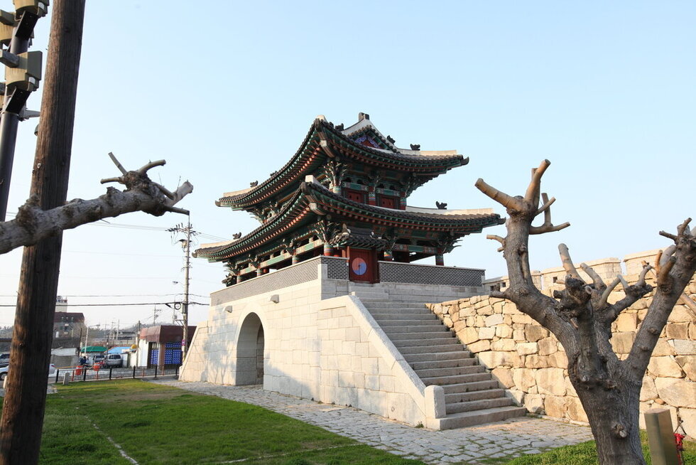 동점문 측면을 찍은 사진. 2층 기와로 되어 있고 대리석 계단 밑으로 입구가 보임. 여러가지 바위로 성벽이 쌓여 있고 그 주위로 나무들이 보임.