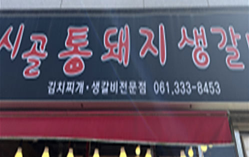 까만색 바탕의 간판에 빨간색 글씨로 시골통돼지생갈비가 크게 적혀 있고 하단에 김치찌개·생갈비전문점 061.333-8453이 하얀색 글씨로 써져있다.