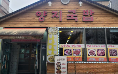 나무 외벽으로 된 양지국밥 식당 외부 전경. 붉은 벽돌과 목재로 된 건물에 양지국밥 간판이 걸려 있고, 출입문 옆 유리창에는 갈비탕, 곱창구이, 양지국밥 등 메뉴 사진과 안내 문구가 붙어 있다. 식당 앞에는 메뉴 입간판이 세워져 있다.