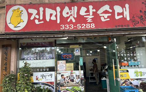 진미옛날순대 식당 외부 전경. 붉은색 간판에 노란 돼지 일러스트와 진미옛날순대 상호, 전화번호 333-5288이 적혀 있다. 출입문 유리에는 영업 중 안내문과 메뉴 사진이 부착되어 있으며, 내부에는 순대국밥 등 음식을 먹는 손님들의 모습이 보인다.