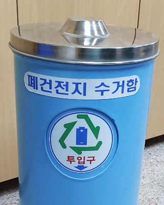 파란색 몸통에 철제로 된 뚜껑으로 되어있고, 정면에는 폐건전지 수거함이란 문구위 아래엔 원형의 투입구가 있는 수거함 모습