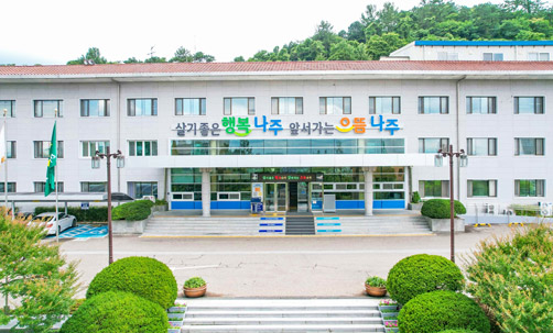 붉은지붕아래 회색빛 3층건물로 청사 정문입구로 올라가는 계단이 있는 청사전경(청사입구에는 살기 좋은 행복 나주 앞서가는 으뜸 나주 슬로건 간판이 걸려있는 모습)