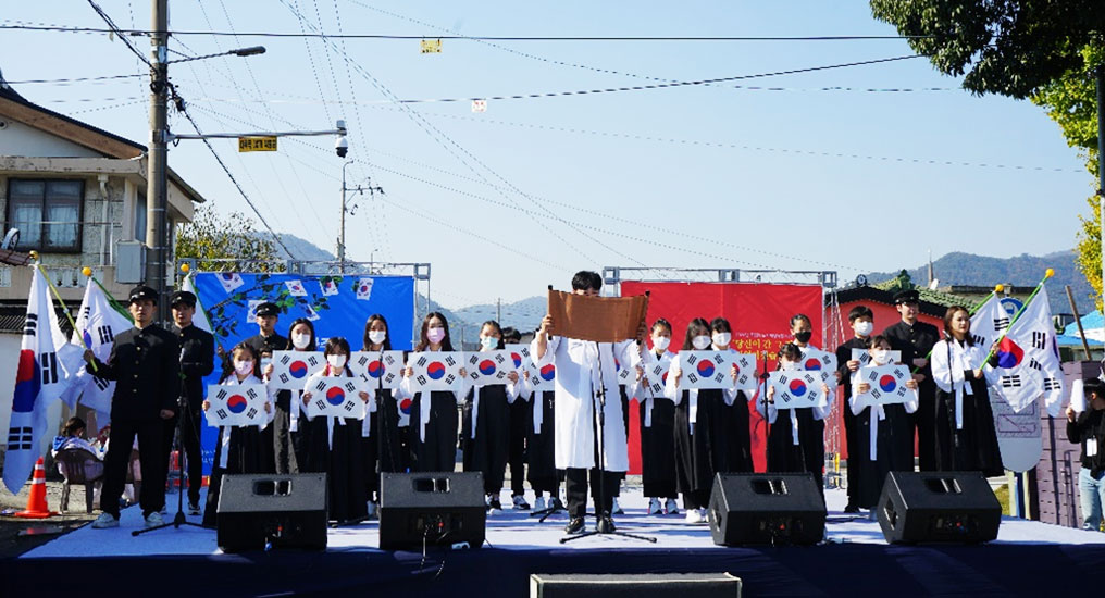 파란하늘 아래 무대가 있고 무대위 뒷쪽에는 학생들이 흰저고리에 검은색치마를 입고 태극기를 들고 있으며 앞쪽 한가운데 남학생이 흰색한복을 입고 말하는 모습