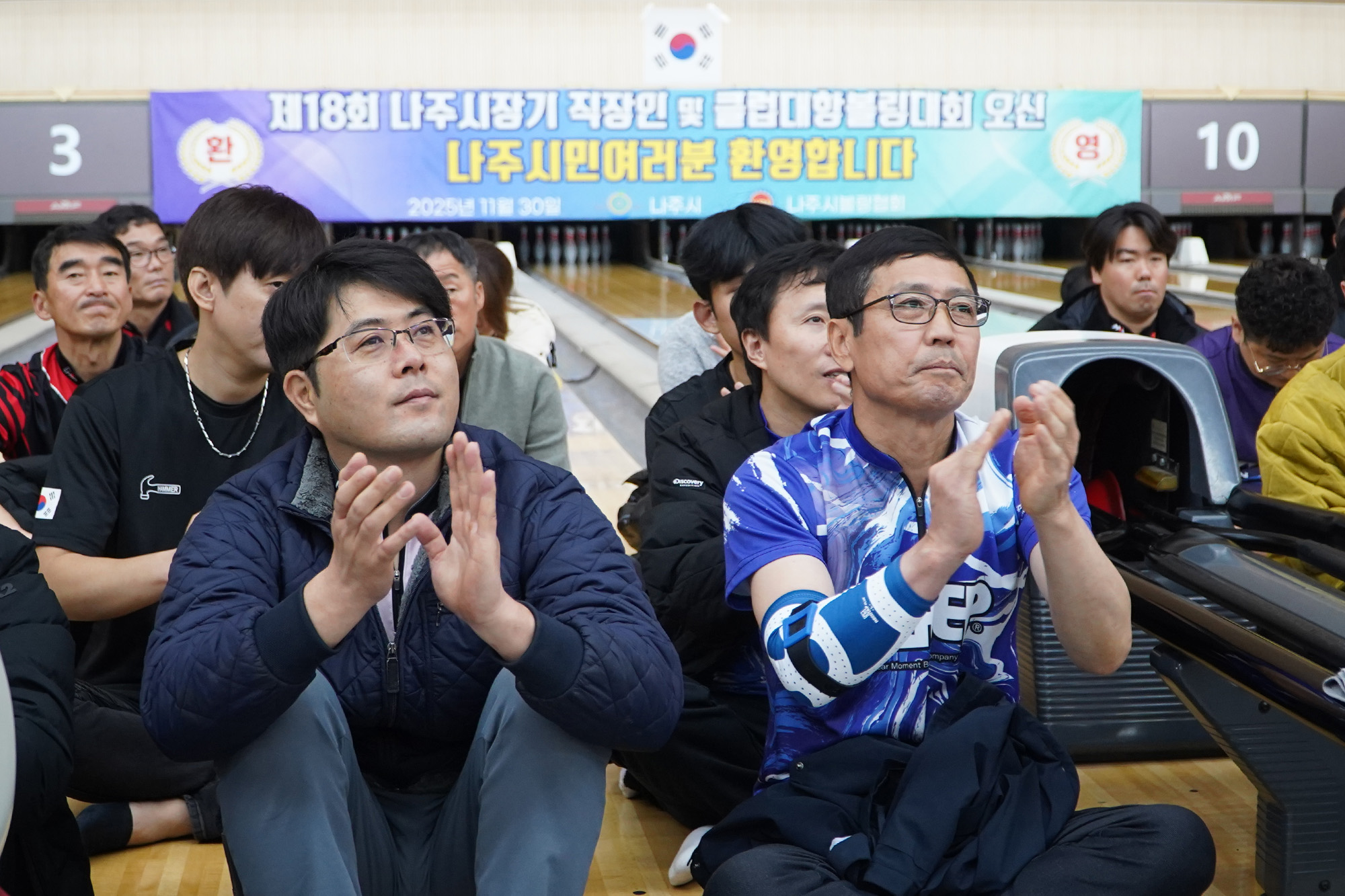  볼링 레인 앞에서 많은 사람들이 앉아 볼링 경기를 관람하고 있으며, 앞줄의 두 남성이 박수를 치고 있다. 배경에는 '제18회 나주시장기 직장인 및 클럽대항볼링대회 오신 나주시민여러분 환영합니다'라고 적힌 현수막이 걸려있다.