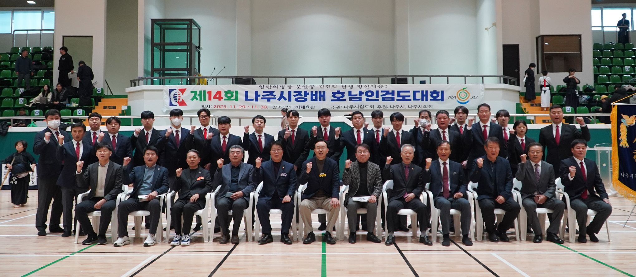  실내 체육관에서 검도 대회에 참석한 관계자들이 단체 사진을 찍는 모습. 앞줄은 의자에 앉아있고 뒷줄은 서 있으며, 많은 사람들이 주먹을 쥐고 파이팅 포즈를 취하고 있다.