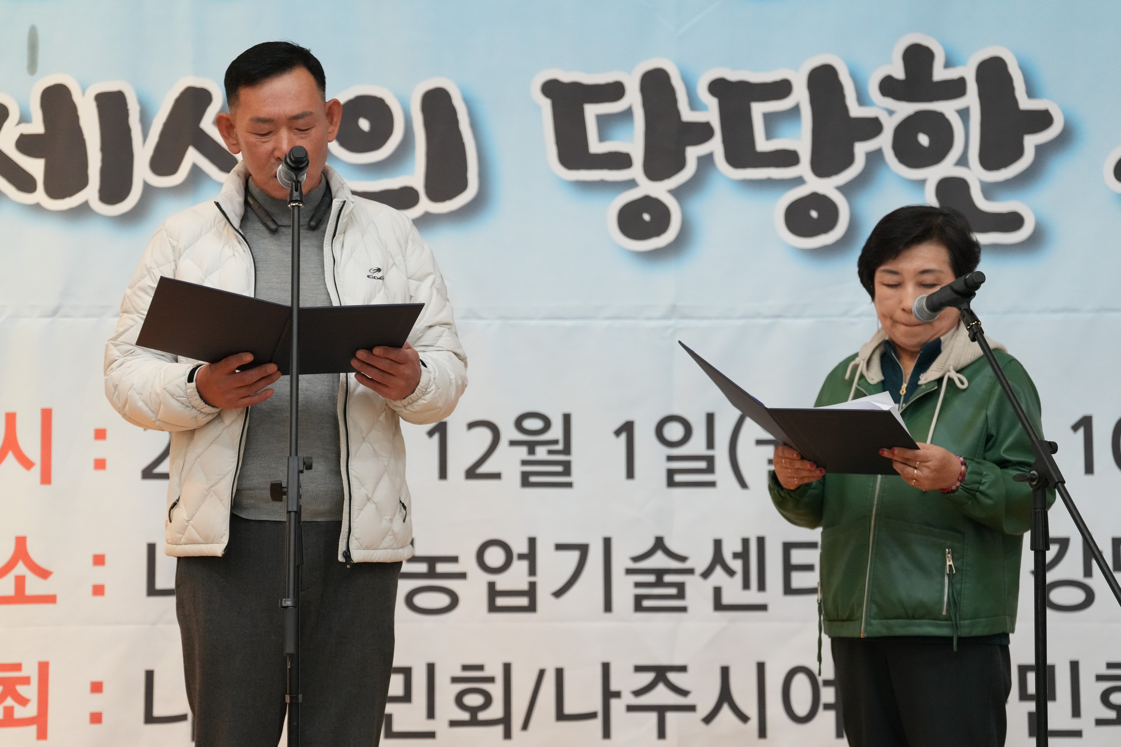 '나주농민 추수대동한마당' 무대 위에서 흰색 패딩을 입은 남성과 녹색 외투를 입은 여성이 각자 자료를 보며 마이크 앞에서 순서를 진행하고 있는 모습
