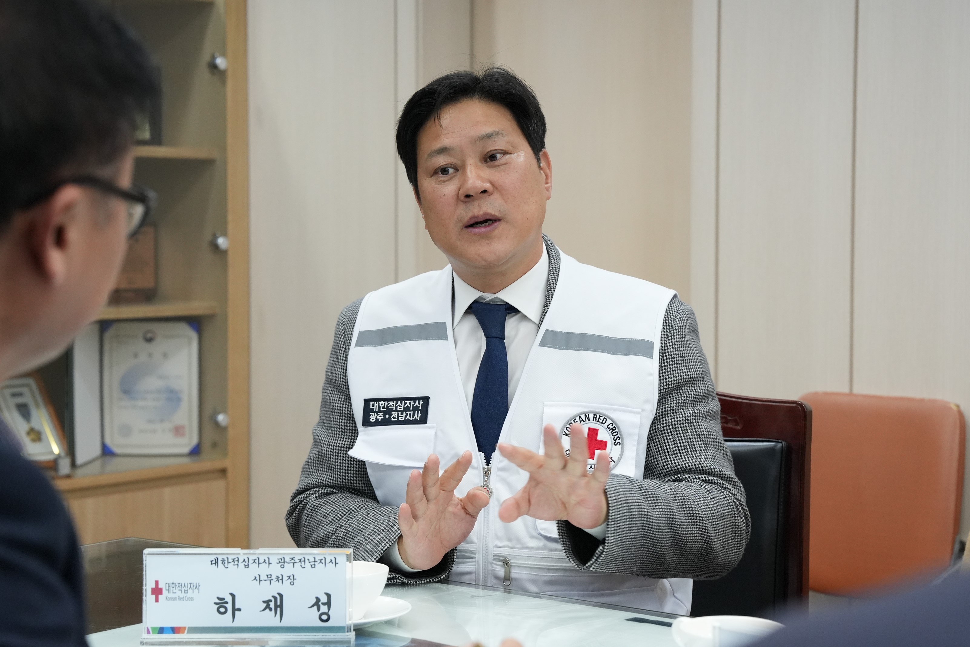  흰색 구호 조끼를 입은 중년 남성이 테이블 맞은편 사람에게 손을 사용해 설명하며 이야기하고 있는 모습