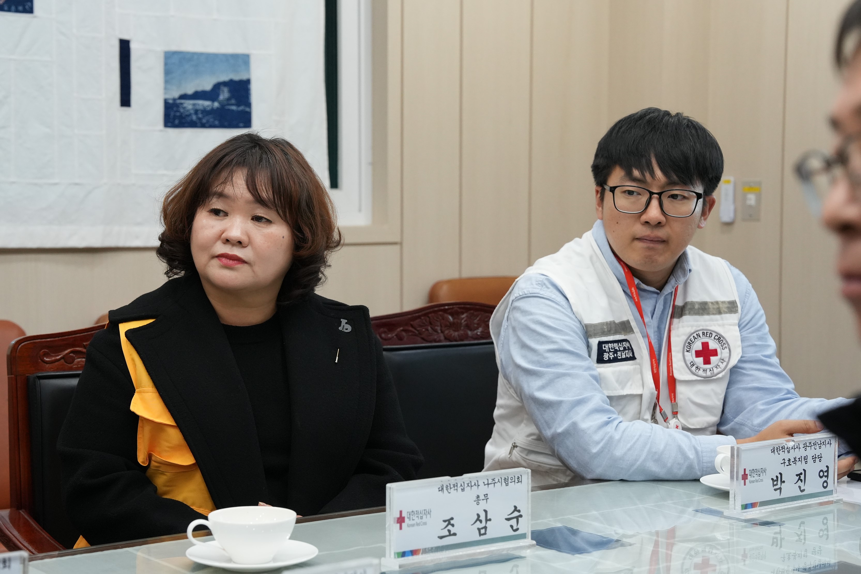  회의 테이블에 나란히 앉아 있는 두 참석자. 왼쪽의 여성은 검은색 외투를 입었고, 오른쪽의 남성은 흰색 조끼를 입고 안경을 쓰고 있다.