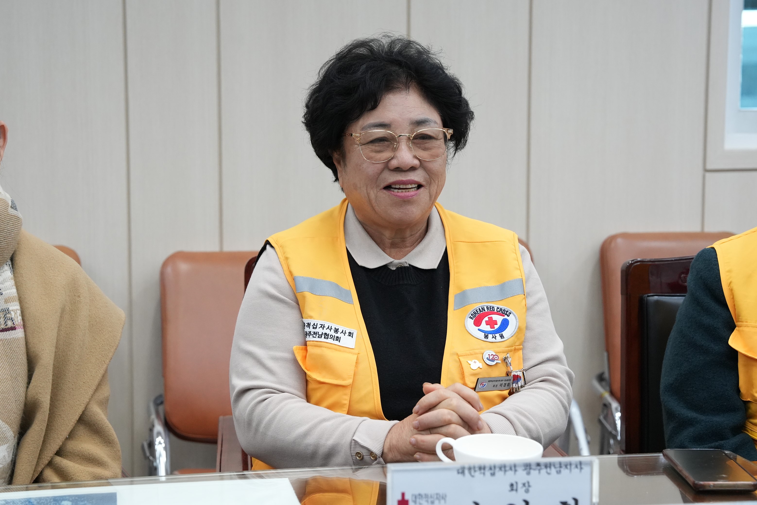  적십자 노란색 조끼를 입은 중년 여성이 테이블에 앉아 밝게 미소 짓고 있는 상반신 클로즈업 사진