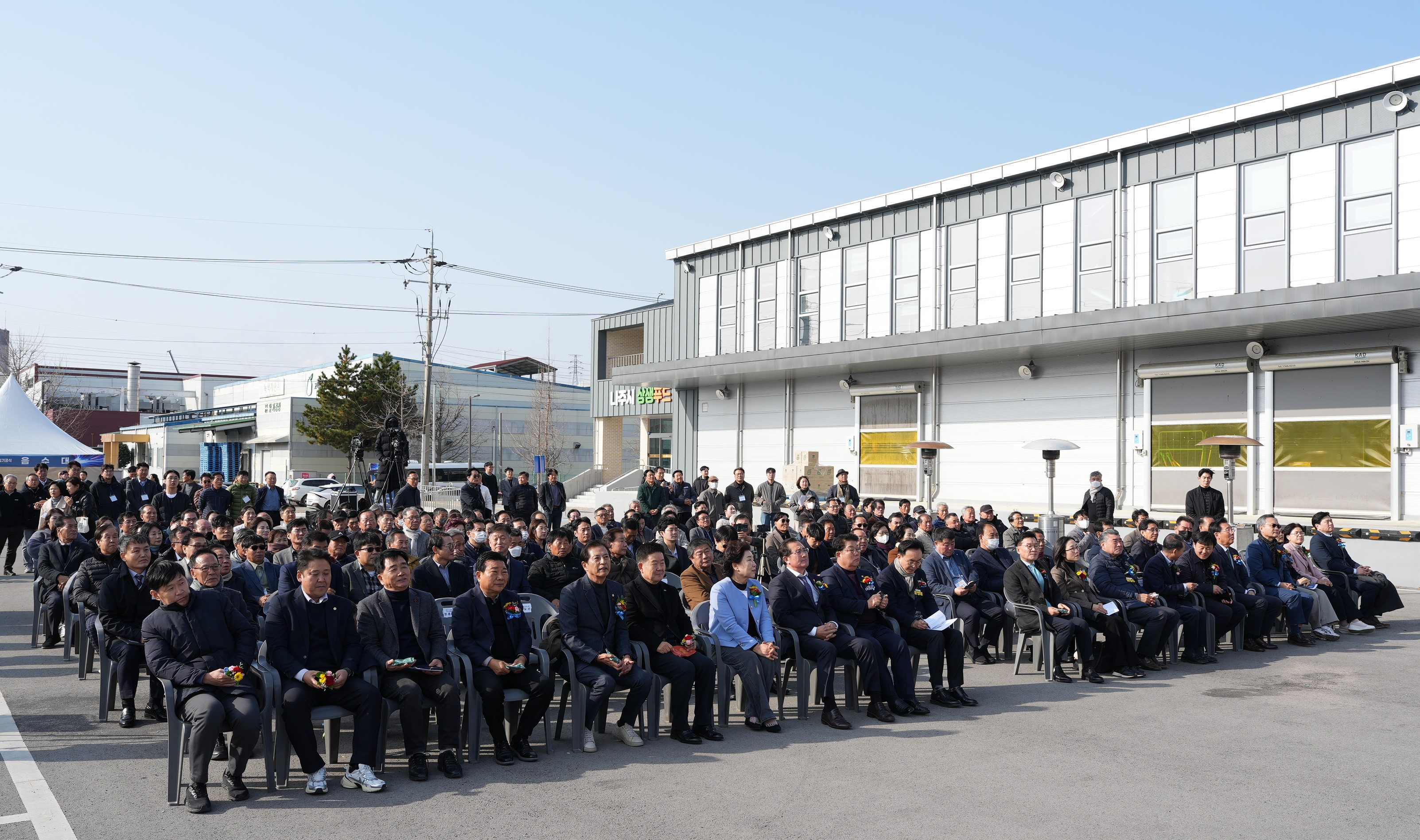  푸드테크 연구지원센터 건립공사 기공식 참석자 전경. 야외 공터에서 많은 참석자들이 일렬로 놓인 의자에 앉아 앞쪽 무대를 향해 경청하고 있다. 뒤쪽으로는 공장 또는 창고 건물들이 보인다.