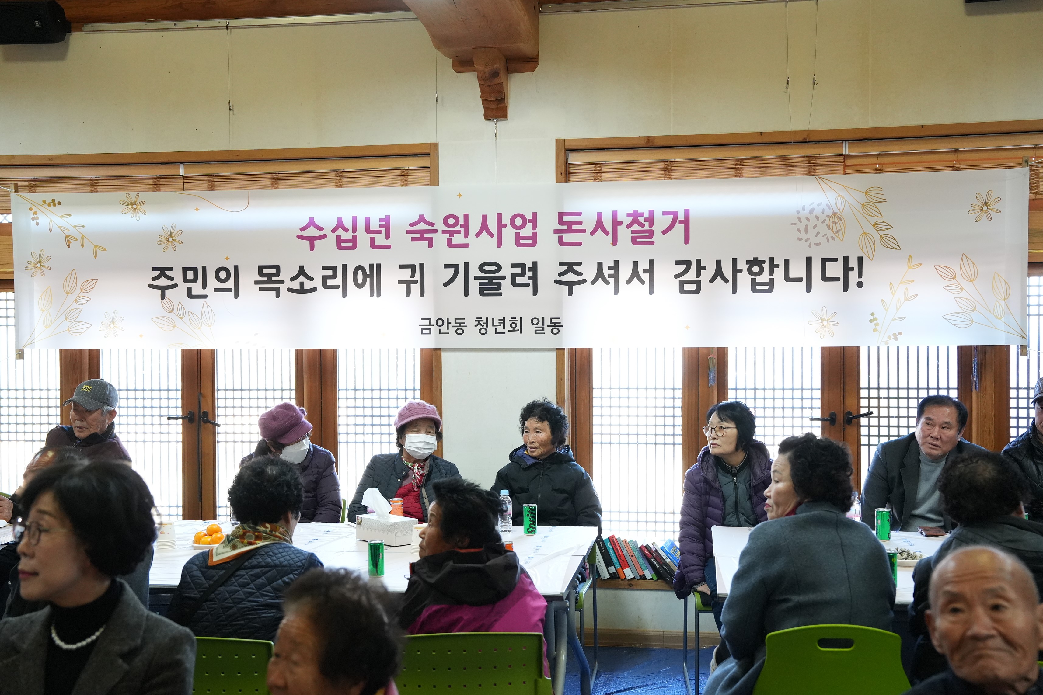  실내 행사에 참석한 주민들의 모습과 현수막. 테이블에 둘러앉은 주민들이 이야기를 나누고 있으며, 뒤쪽 창문 위에 '수십년 숙원사업 돈사 철거, 주민의 목소리에 귀 기울여 주셔서 감사합니다! 금안동 청년회 일동'이라고 적힌 현수막이 걸려 있다.