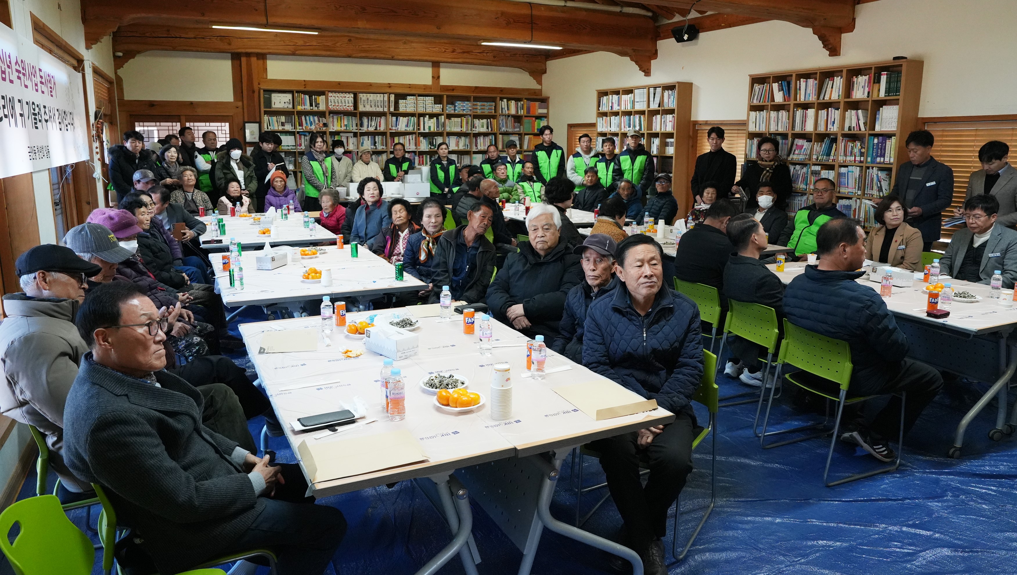  금안마을 행사장의 넓은 전경. 테이블에 둘러앉은 많은 마을 주민들이 앞쪽 무대를 향해 앉아 행사에 참여하고 있다. 실내 양쪽 벽면은 책장으로 가득 차 있다.