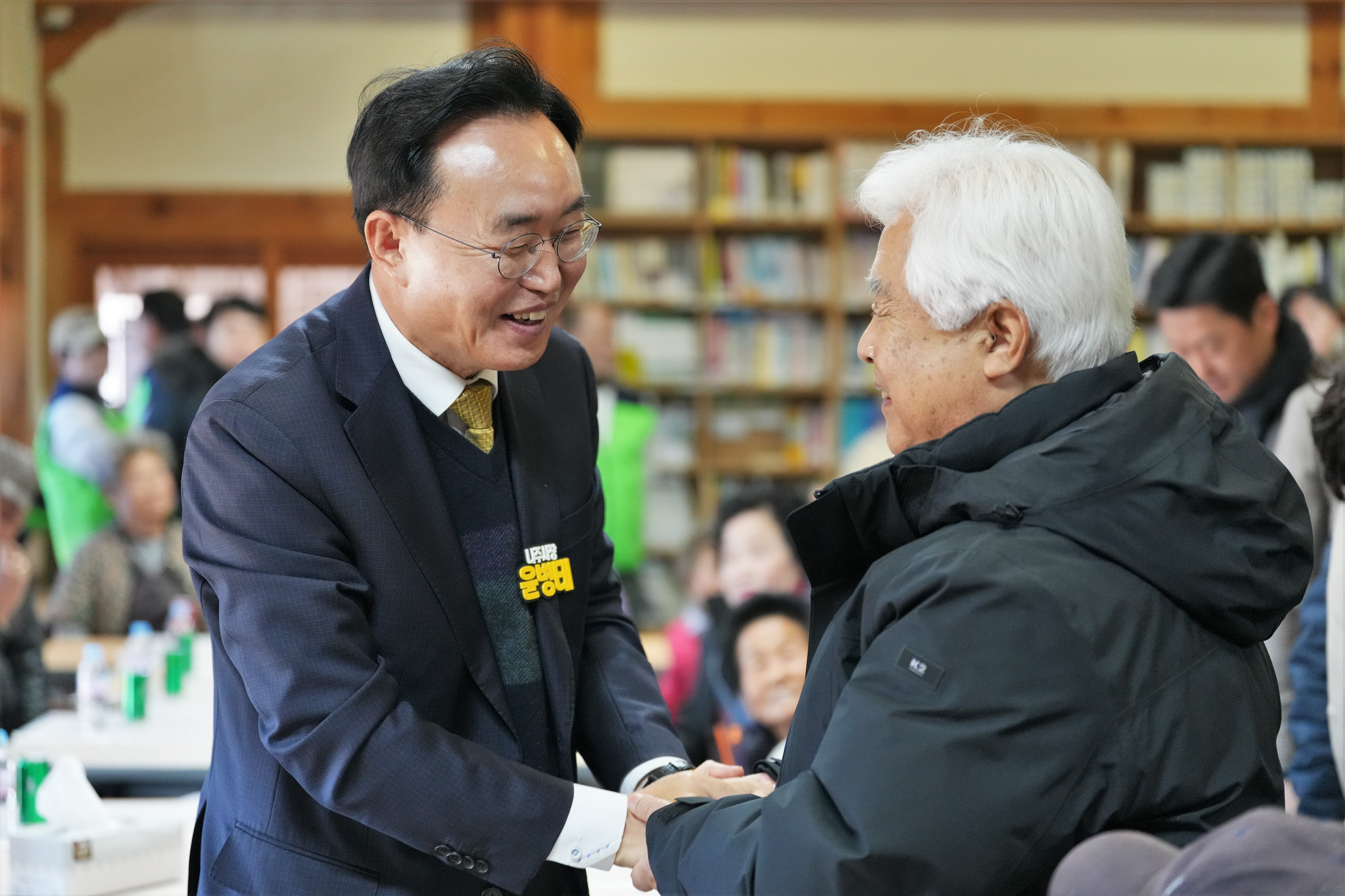  윤병태 나주시장이 한 노년 남성과 악수하며 환하게 웃는 모습. 주변에는 많은 참석자들이 테이블에 둘러앉아 있다.