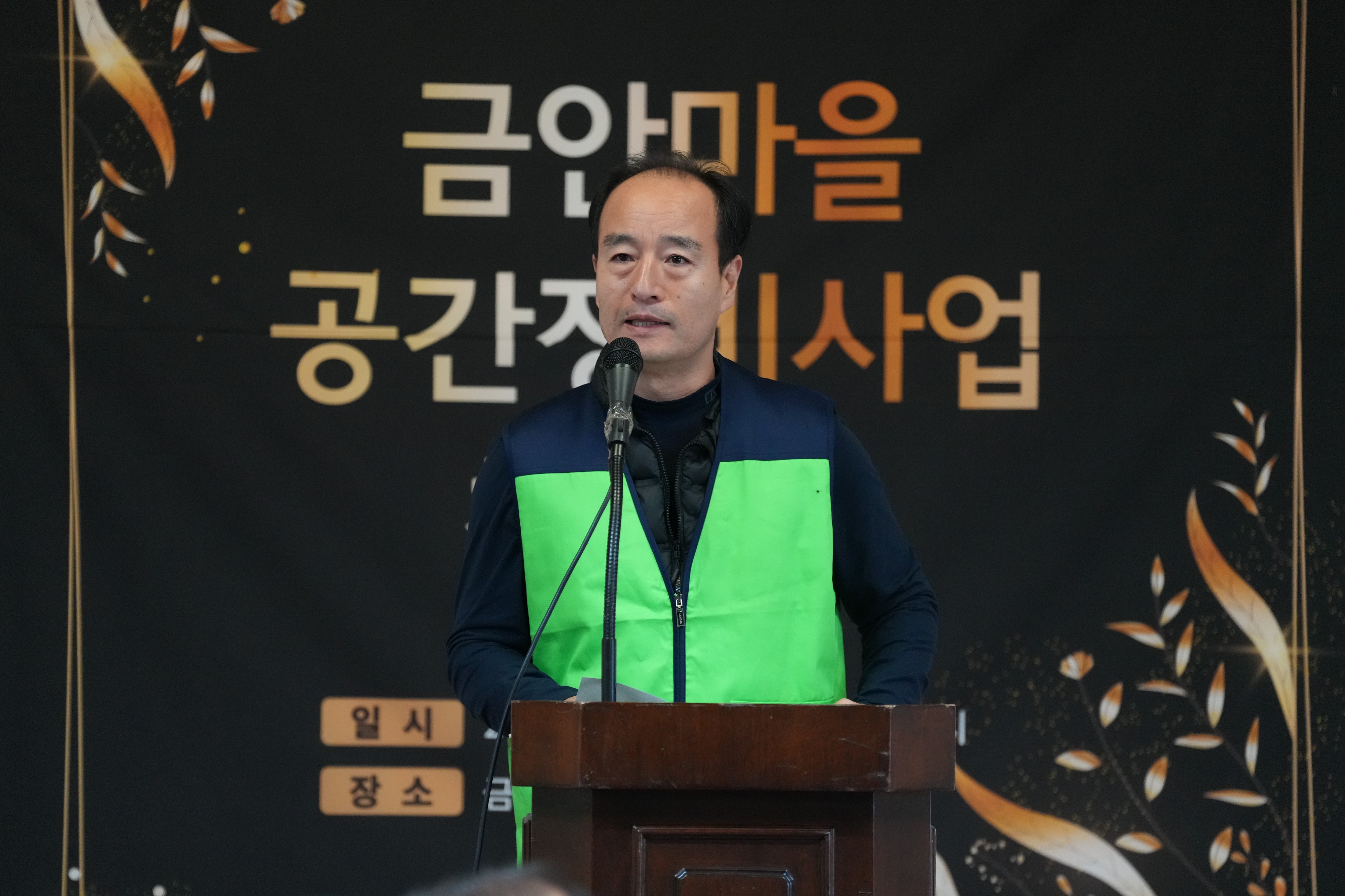  녹색 조끼를 입은 중년 남성이 단상에서 발언하는 모습. 배경에는 '금안마을 공간재생사업' 현수막이 보인다.