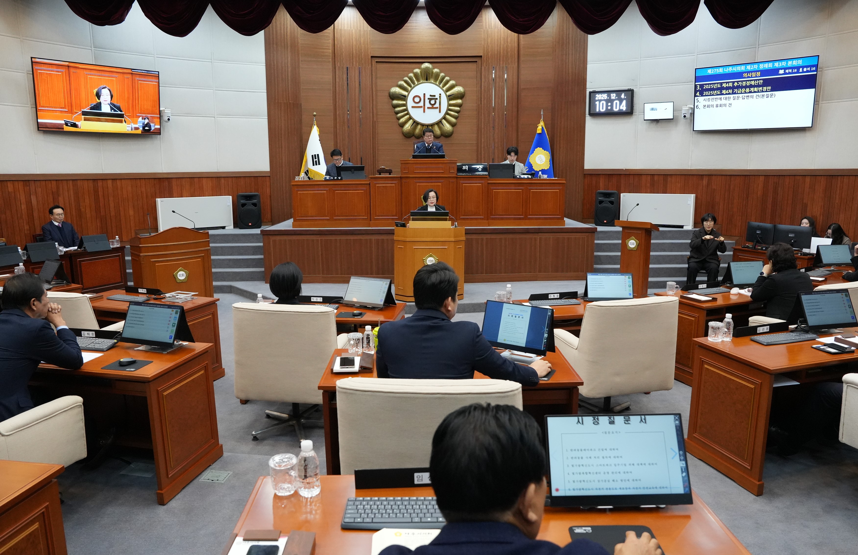  시의회 본회의장 전경. 의원들이 책상에 앉아 있고, 중앙 단상에서 발언하는 사람이 있으며, 벽면 스크린에는 '의회' 글자와 회의 내용이 보임