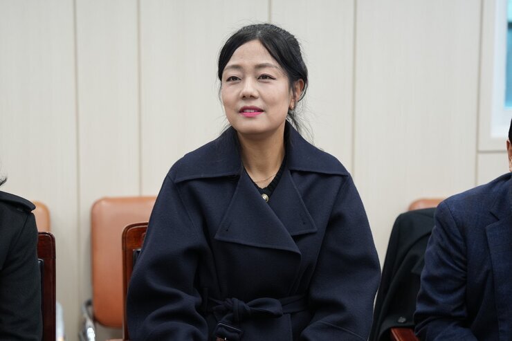 회의석상에 앉아 미소를 짓고 있는 중년 여성의 클로즈업 사진. 진한 남색 코트를 입고 있다.