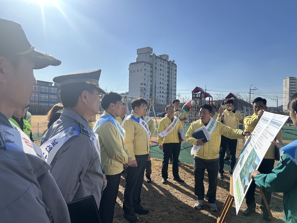 맑은 날 야외에서 여러 사람이 모여 서류를 보며 대화하고 있다. 장소는 놀이터나 공터로 보이며, 뒤편에는 아파트와 학교 건물이 보인다. 모인 사람들 중 상당수는 노란색 점퍼를 입고 있으며, '순찰'이나 '지역 방범'과 관련된 것으로 보이는 어깨띠를 두르고 있다. 앞쪽 중앙의 노란색 점퍼를 입은 한 남성이 들고 있는 큰 도표나 계획표를 가리키며 설명하고 있다.