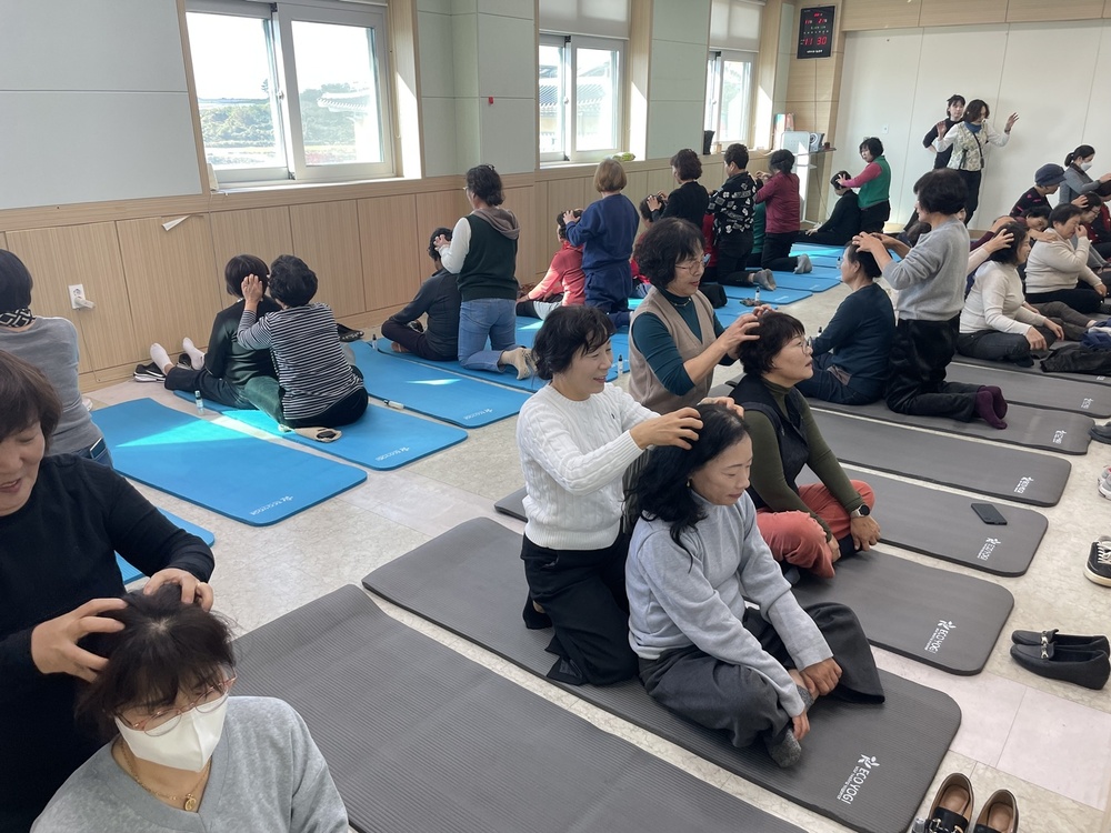 실내 체육 활동 공간에서 중년 여성들이 요가 매트 위에 앉아서 서로의 머리를 마사지 해주는 모습. 모두 편안한 복장이며, 밝고 건강한 분위기이다.