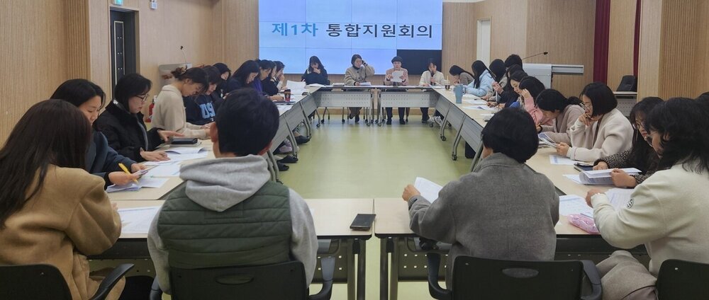 긴 테이블에 둘러앉아 서류를 보며 회의를 하고 있는 여러 사람들의 모습. 벽에 걸린 스크린에는 '제1차 통합지원회의' 라는 글자가 보인다.