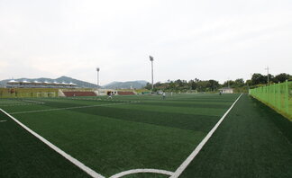 인조잔디구장을 코너에서 찍은 사진. 초록색 인조잔디위에 축구경기를 하고 있는 사진.