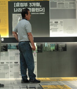 노무현 전대통령 사진과 투표율 70.2% 역대 대선 최저, 무정부 3개월 나라가 흔들린다(2003.5.20. 조선일보) 등 여러 신문시가사 붙혀져있는 전시장을 보고있는 관람객 둘