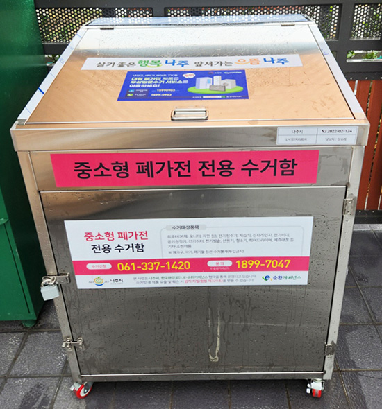 정사각의 철제 수거함으로 뚜껑엔 나주로고, 정면엔 진분홍배경에 까만 글씨로 중소형 폐가전 전용 수거함이라 적혀있고 그 밑에는 061-337-1420, 1899-7047, 나주시 로고가 적혀있고 옆에는 자물쇠로 고리가 잠겨있는 모습