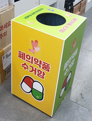노란색과 초록색으로 면마다 다르게 구성된 윗부분에 원형으로 구멍이 나있는 길죽한 몸통의 폐의약품 수거함이라고 적힌 수거함 모습