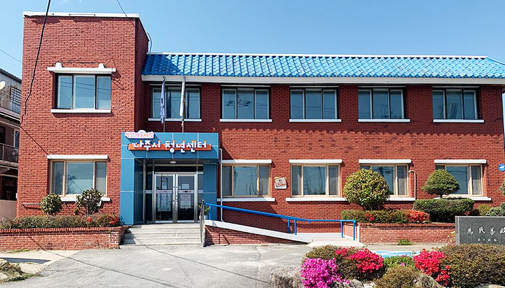 파란지붕에 벽돌로 지어진 나주시 청년센터 건물이 있고 건물 앞에 꽃들이 피어 있다.