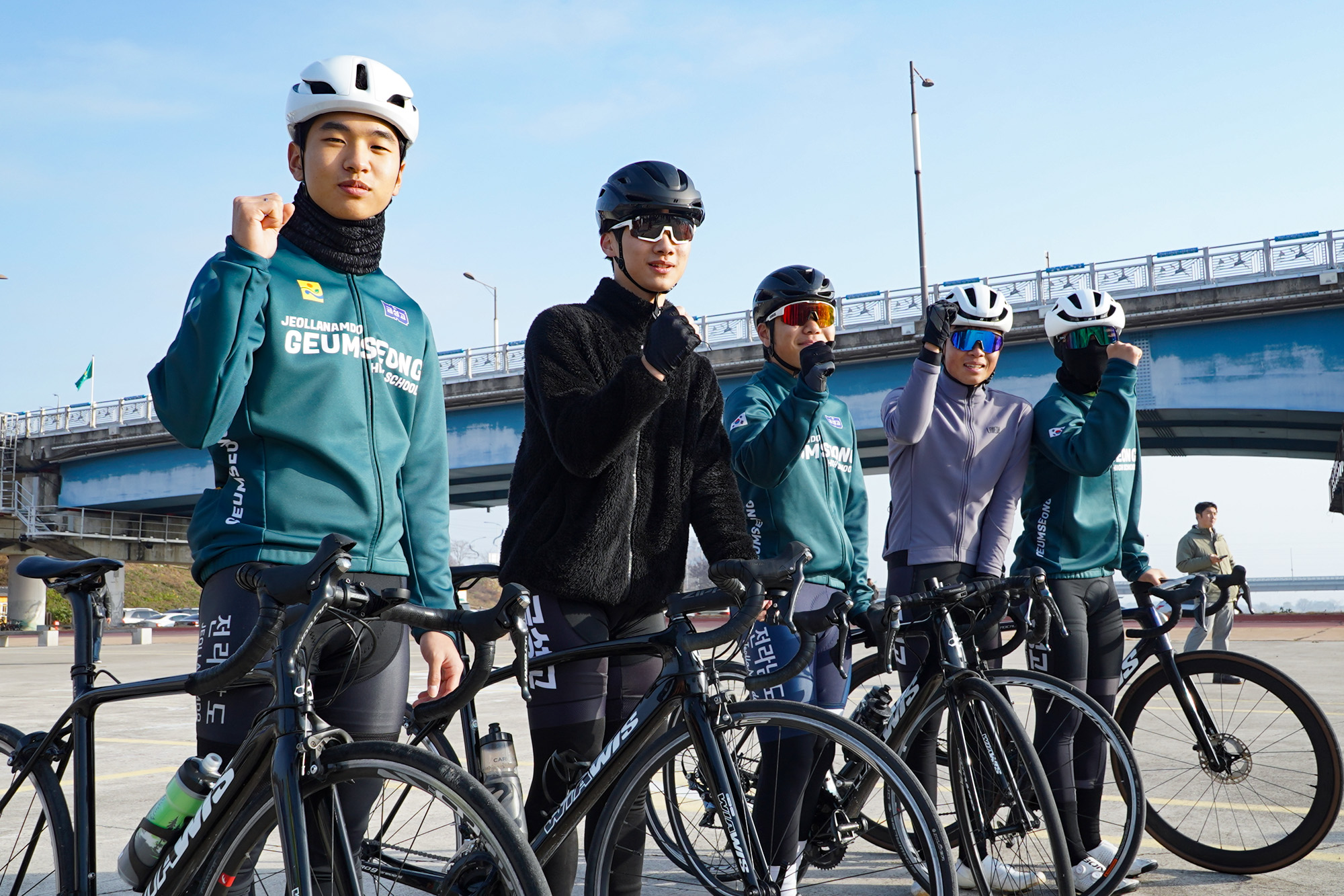 자전거와 헬멧을 착용한 젊은 사이클 선수 네 명이 야외 다리 아래에서 각자 주먹을 쥐거나 포즈를 취하며 정면을 바라보고 있다.