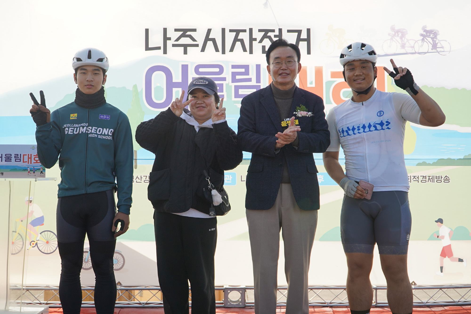 '나주시자전거 어울림대회' 현수막 앞에서 윤병태 나주시장이 자전거 복장을 한 남성 두 명과 캐주얼 복장의 여성 한 명과 함께 서서 포즈를 취하고 있다. 참가자들은 'V' 표시를 하고 있다.