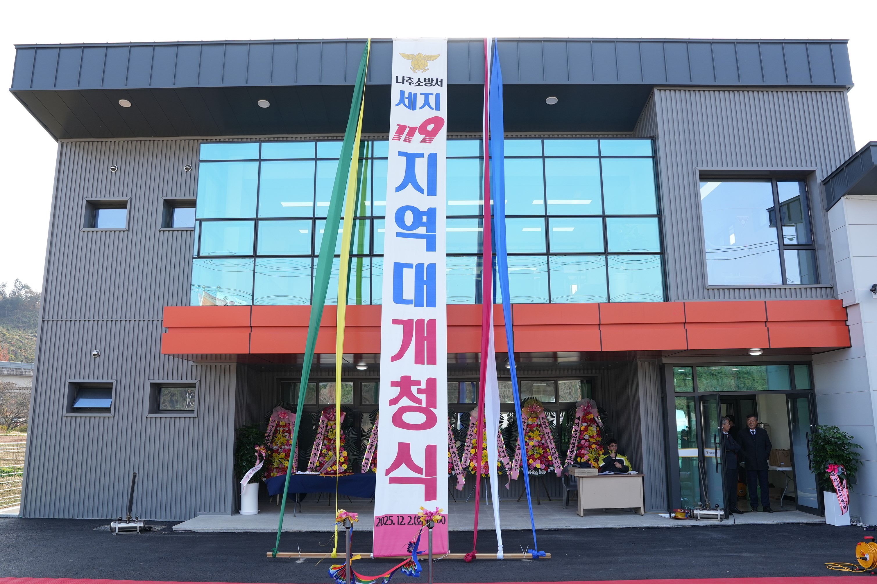  신축된 나주소방서 세지 119 지역대 건물 외관. 깔끔한 회색 외벽에 주황색 포인트가 들어간 2층 건물이며, 정면에는 세로로 길게 '나주소방서 세지 119 지역대 개청식' 현수막이 걸려있다. 건물 주변에는 화환들이 놓여있다.