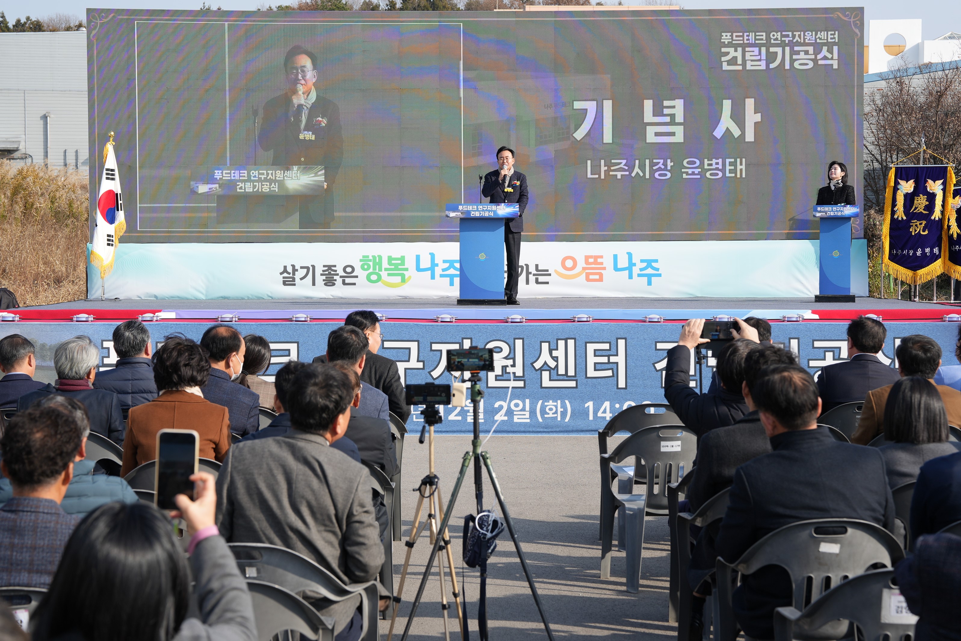  푸드테크 연구지원센터 건립기공식 행사 전경. 무대 중앙에 윤병태 나주시장이 연설하고 있으며, 대형 스크린에 그의 모습과 '기념사 나주시장 윤병태'라는 문구가 표시되어 있다. 참석자들이 의자에 앉아 행사를 관람하고 있으며, 일부는 스마트폰으로 사진을 찍고 있다.