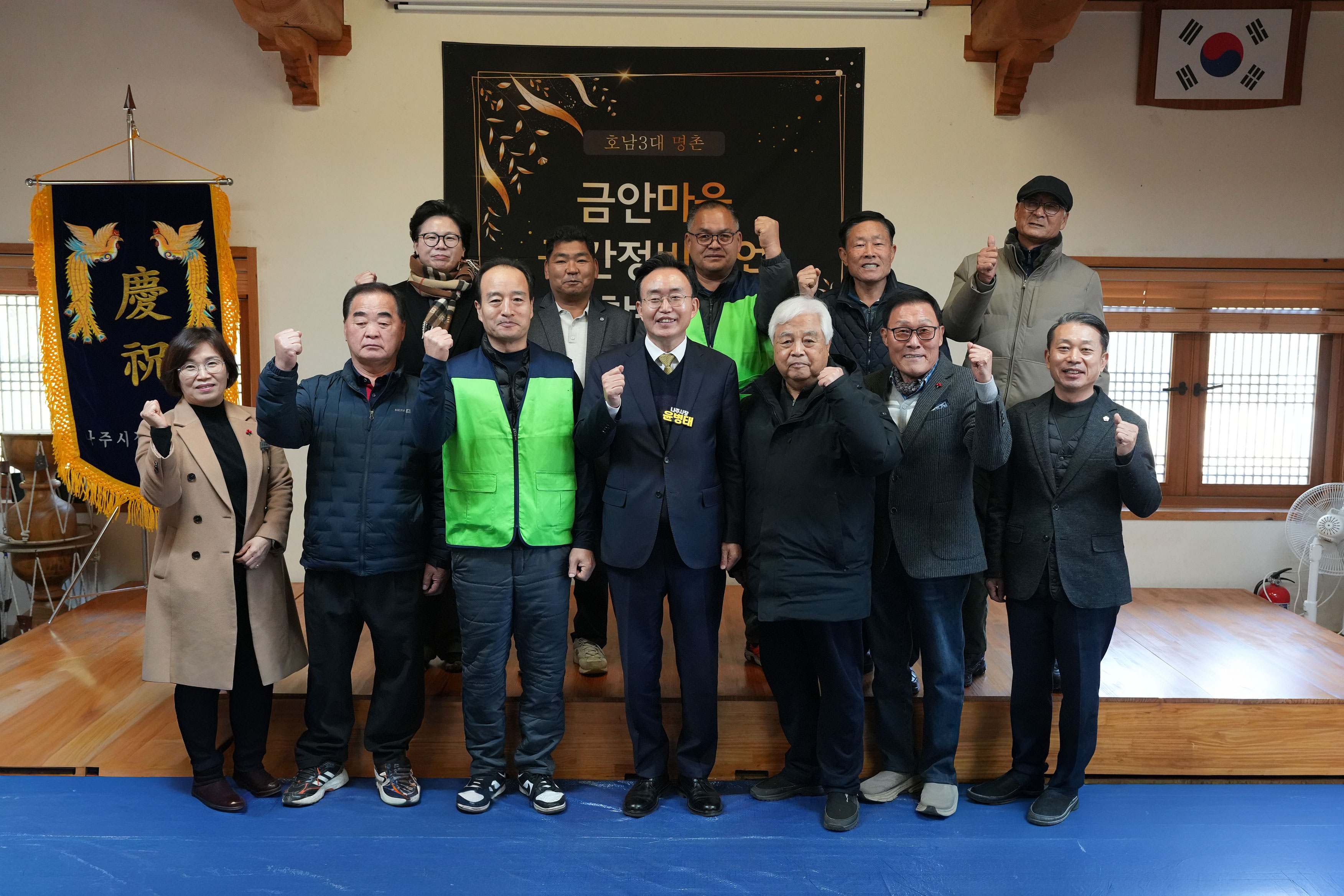  '금안마을 공간재생사업' 행사 참석자들이 무대 앞에서 단체 기념 촬영하는 모습. 10여 명의 남성과 여성이 카메라를 향해 주먹을 쥐거나 엄지손가락을 들며 포즈를 취하고 있다. 중앙의 남성(나주시장 윤병태)은 정장 차림이며, 일부 남성은 녹색 조끼를 착용하고 있다.