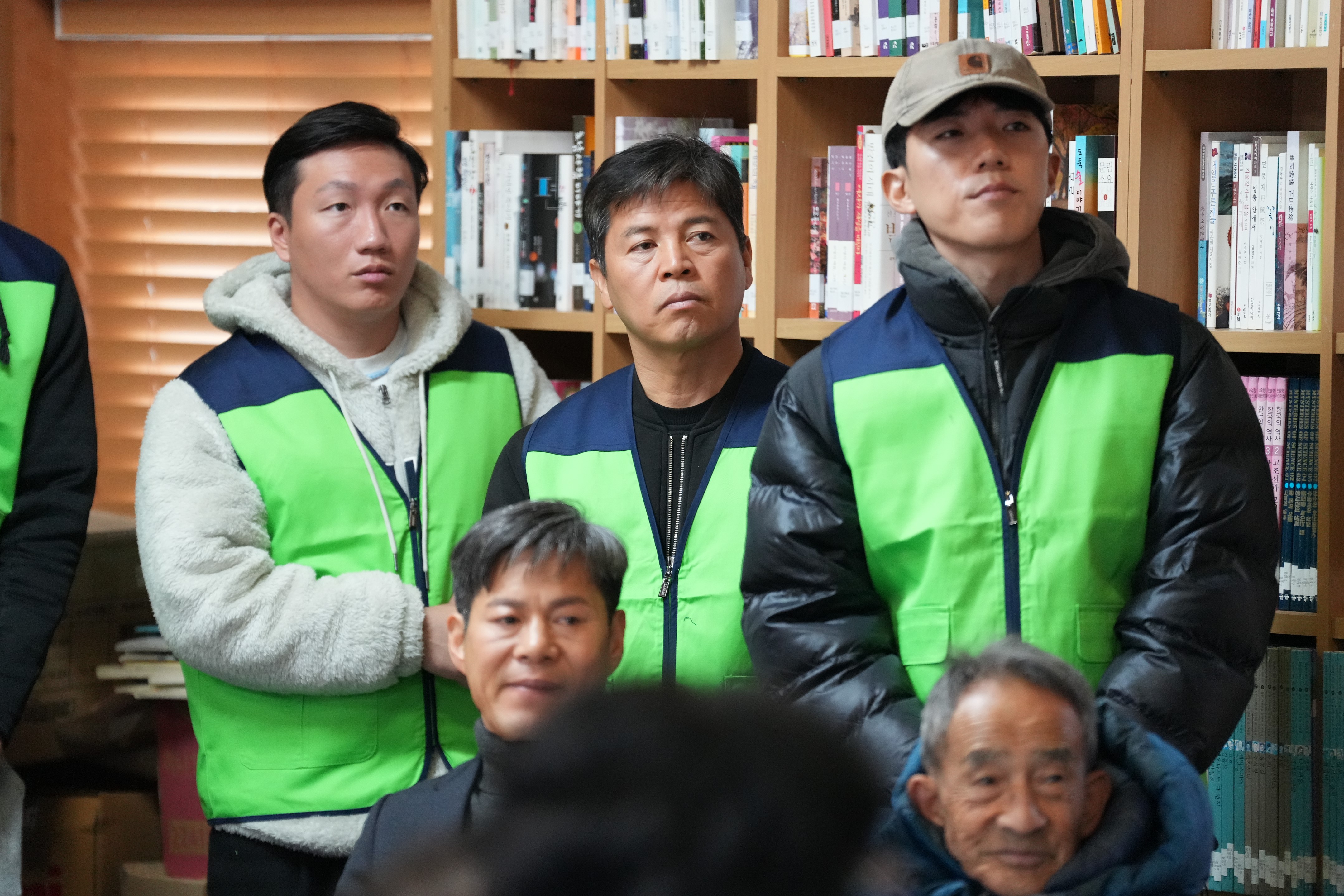  녹색과 파란색 조끼를 입은 세 명의 남성 등 참석자들이 경청하는 모습의 클로즈업 사진. 배경에는 책들이 가득한 책장이 보인다.