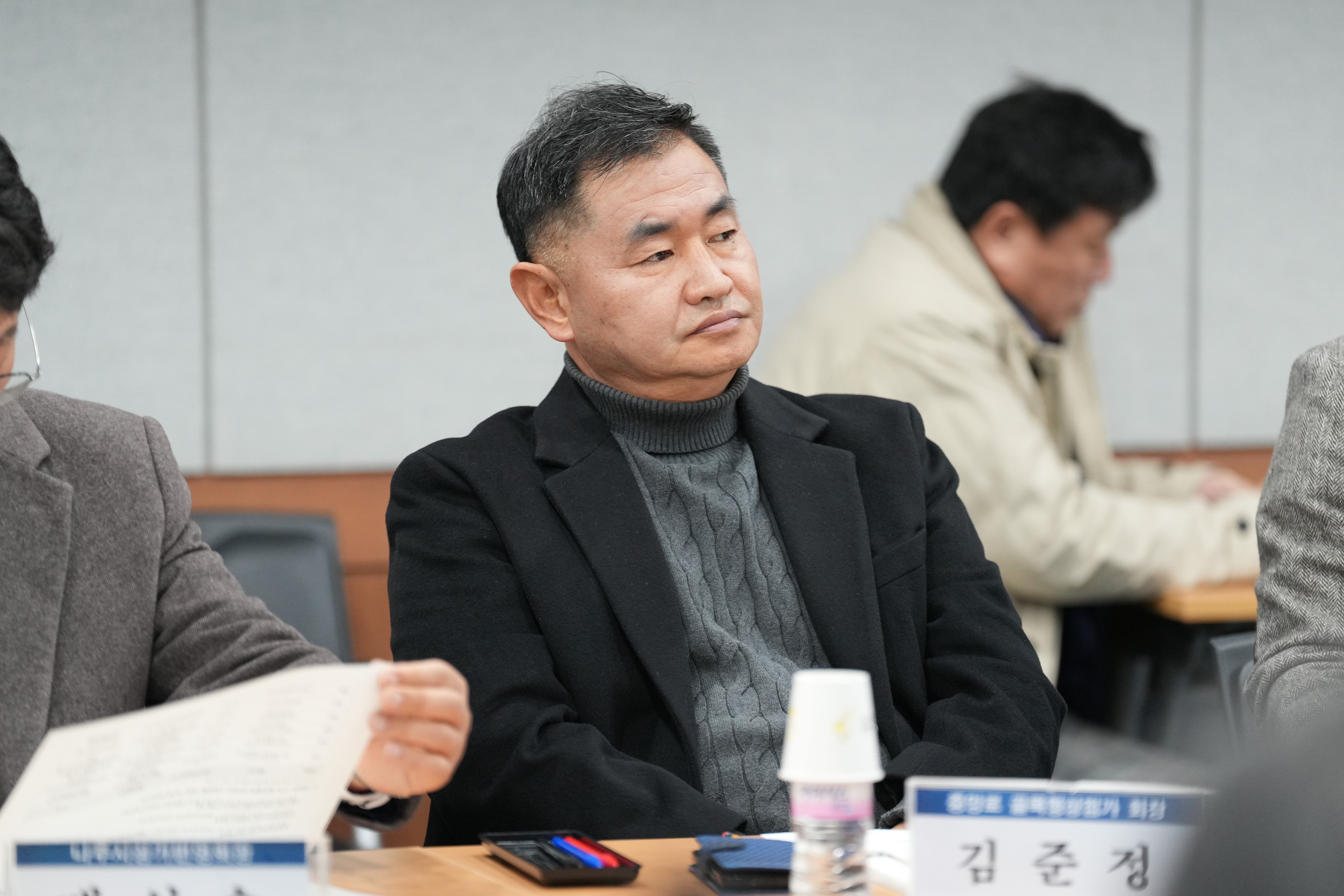 회의석상에 앉아 정면을 응시하는 남성의 모습. 검은색 코트와 회색 터틀넥 스웨터를 입고 있으며, 앞에 '김준정'이라고 적힌 명패가 보인다.