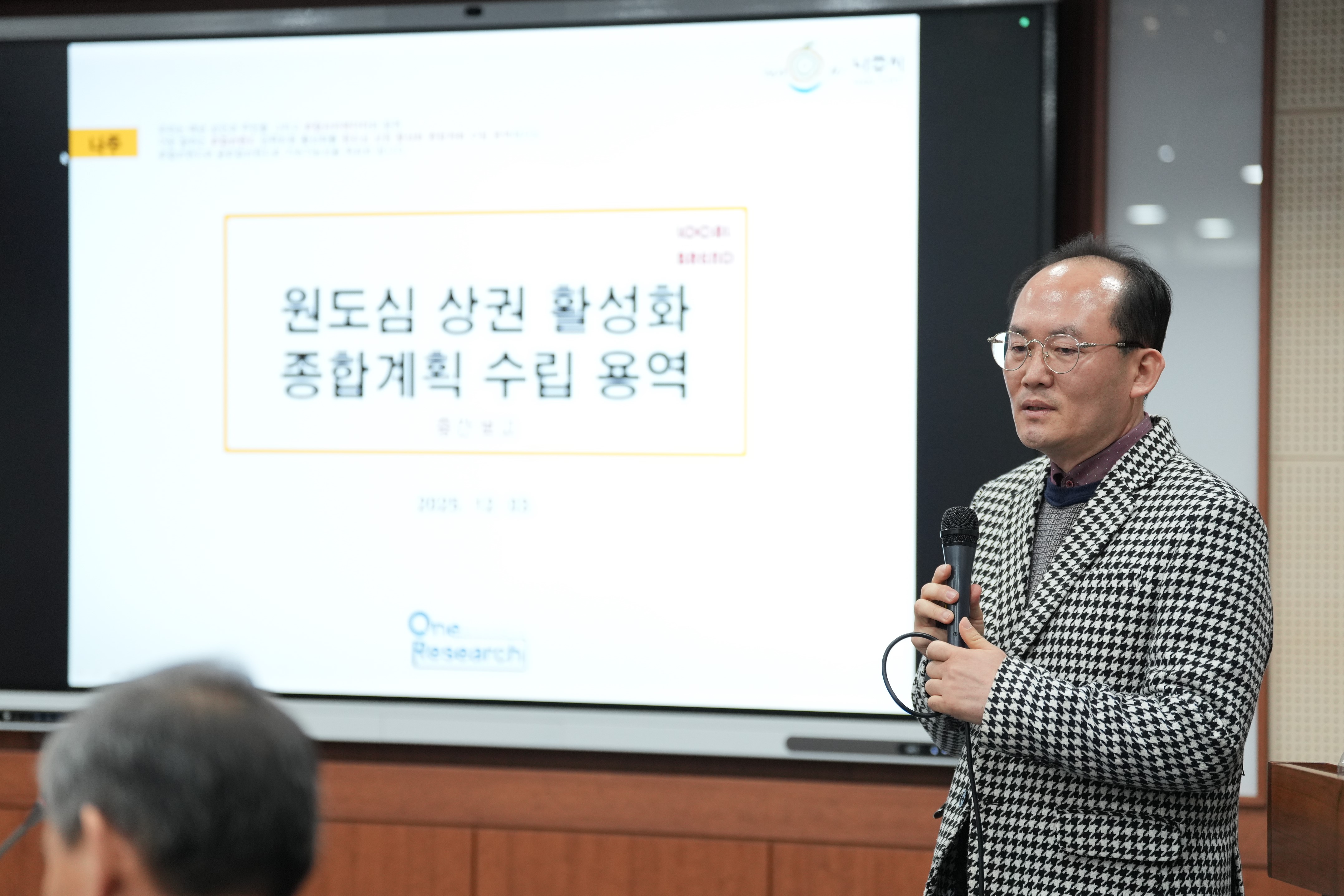 발표를 진행하는 중년 남성. 체크무늬 재킷을 입은 남성이 마이크를 잡고 스크린 앞에서 이야기하고 있다. 스크린에는 '원도심 상권 활성화 종합계획 수립 용역'이라는 제목의 슬라이드가 보인다.