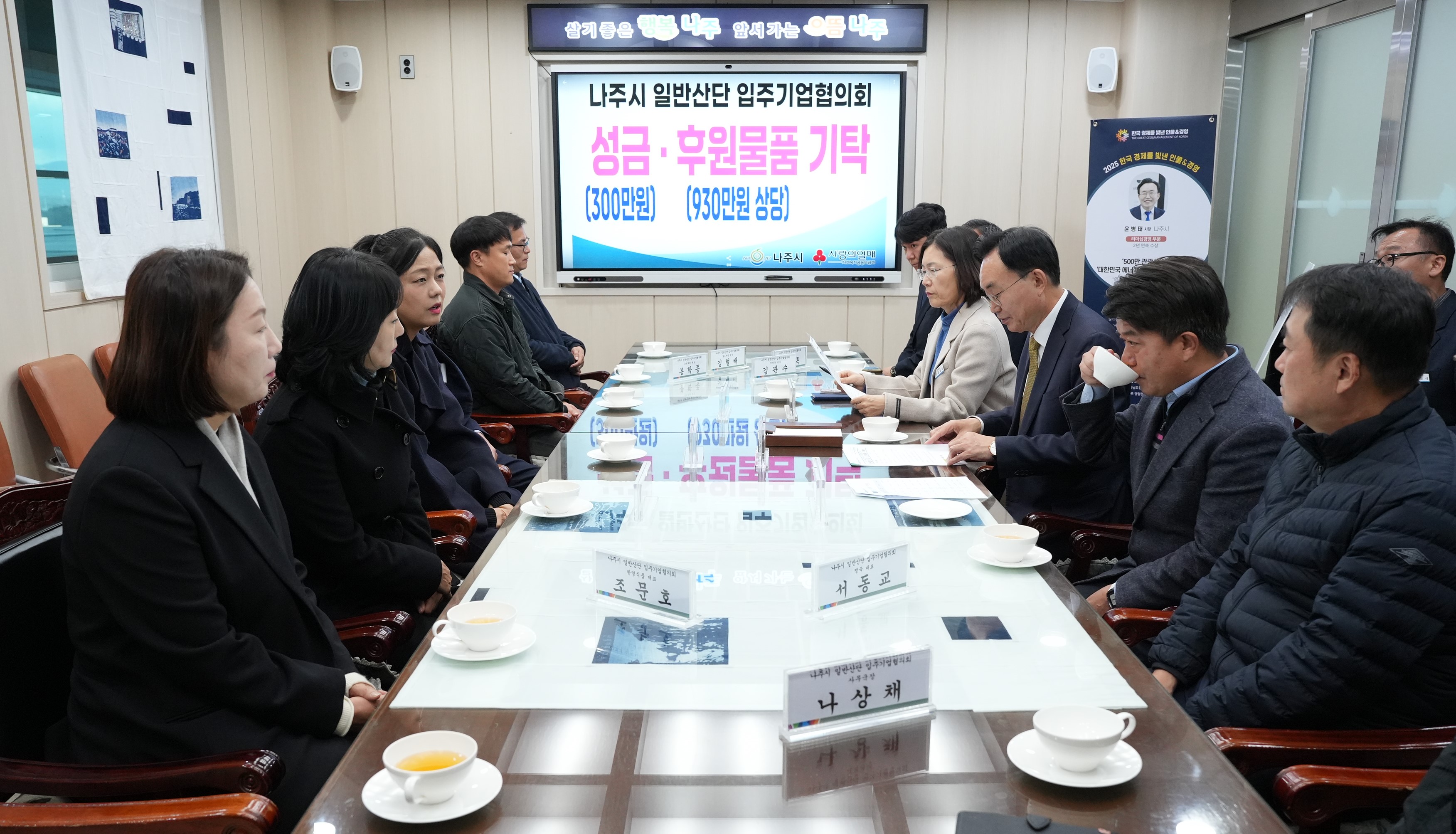  회의 또는 간담회 장면. 직사각형 테이블에 마주 앉은 나주시 관계자들과 기업 관계자들이 대화를 나누고 있다. 테이블 위에는 '조광호', '서영고', '나상채' 등의 명패가 놓여 있으며, 뒤쪽 화면에는 '나주시 일반산단 입주기업협의회 성금·후원물품 기탁 (300만원) (930만원 상당)'이라는 문구가 보인다.