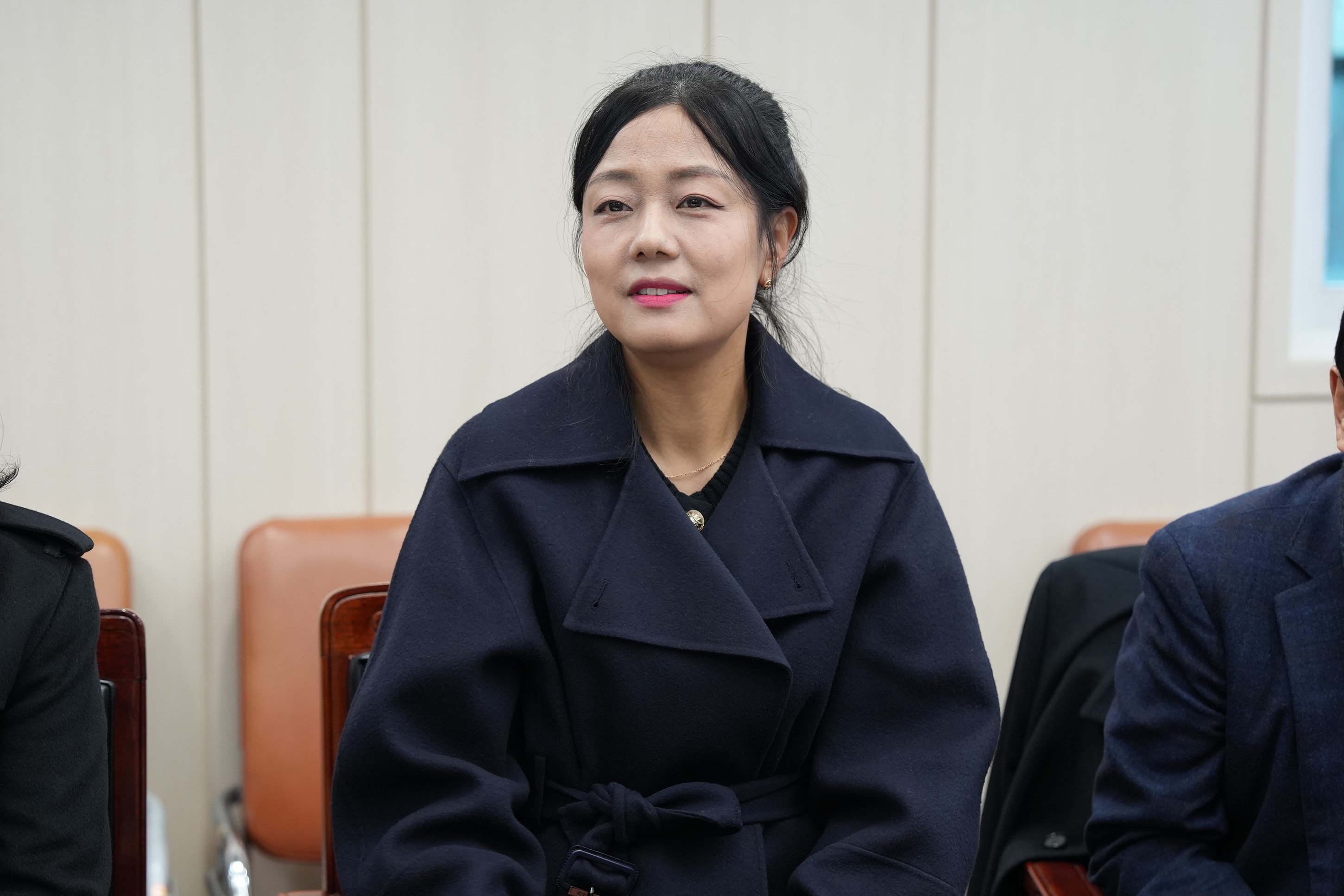  회의석상에 앉아 미소를 짓고 있는 중년 여성의 클로즈업 사진. 진한 남색 코트를 입고 있다.