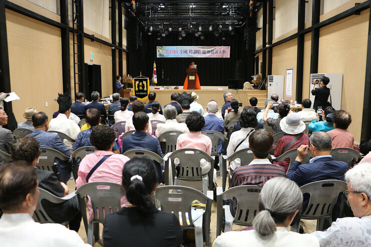 전국 시조창 나주 경연대회 무대 위의 여성 참가자가 자리에 서서 경연을 하고 있는 모습과 참석자들이 앉아서 듣고 있는 뒷모습