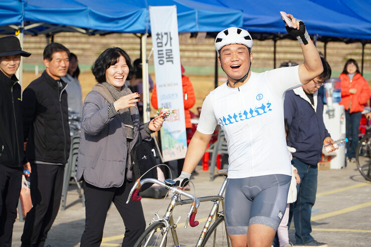  자전거 복장과 헬멧을 착용한 남성 사이클리스트가 자신의 자전거 옆에서 한 손을 들고 환하게 웃고 있으며, 옆의 여성 관람객도 웃으며 박수를 치고 있다.