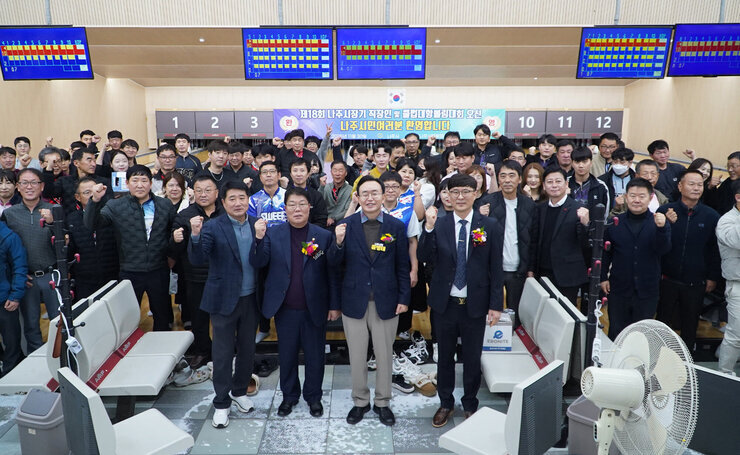 볼링 레인 앞에서 다수의 사람들이 단체 사진을 찍고 있다. 앞줄의 몇몇 주요 인사들이 주먹을 쥐고 포즈를 취하고 있다. 배경에는 볼링 점수판과 대회 현수막이 보인다.