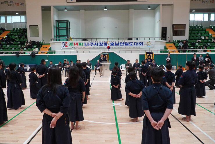  실내 체육관에서 검도 대회 개회식이 진행되는 모습. 검도복을 입은 많은 선수들이 도장 위에 도열해 있고, 단상 위에서는 윤병태 나주시장이 연설하고 있다. 배경에는 '제14회 나주시장배 호남인검도대회'라는 현수막이 걸려있다.
