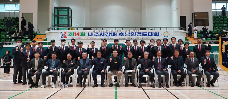  실내 체육관에서 검도 대회에 참석한 관계자들이 단체 사진을 찍는 모습. 앞줄은 의자에 앉아있고 뒷줄은 서 있으며, 많은 사람들이 주먹을 쥐고 파이팅 포즈를 취하고 있다.