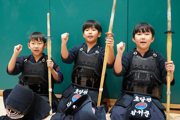  세 명의 어린 남자아이들이 검도 도복과 호구를 착용하고 죽도를 들고 앉아있다. 모두 주먹을 쥐고 파이팅 포즈를 취하고 있으며, 웃는 표정으로 카메라를 바라본다.