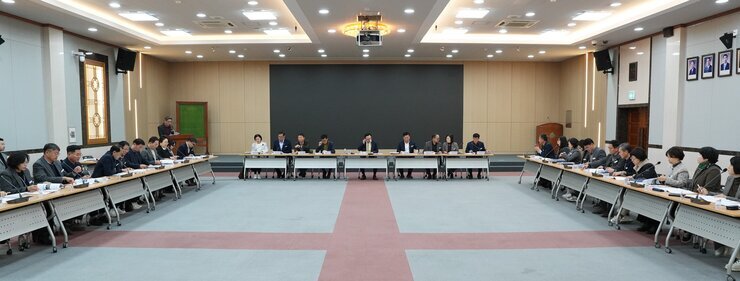 넓은 회의장에서 'ㄷ'자 형태로 테이블에 둘러앉아 회의하는 많은 사람들의 모습. 중앙에는 주요 관계자들이, 양 옆 긴 테이블에는 여러 참석자들이 앉아있다. 뒤쪽 벽에는 검은색 스크린과 액자들이 걸려있다.
