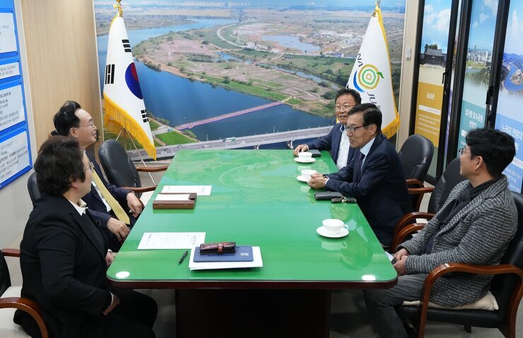 다섯 명의 남성(나주시장 윤병태 포함)이 큰 녹색 테이블에 둘러앉아 회의를 하는 모습. 정장을 입은 사람들이 보이며, 테이블 위에는 서류와 찻잔이 놓여 있다. 뒤쪽 벽에는 태극기와 나주 로고가 있는 깃발, 그리고 강이 보이는 야외 전경 사진이 걸려 있다.