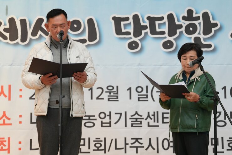 '나주농민 추수대동한마당' 무대 위에서 흰색 패딩을 입은 남성과 녹색 외투를 입은 여성이 각자 자료를 보며 마이크 앞에서 순서를 진행하고 있는 모습